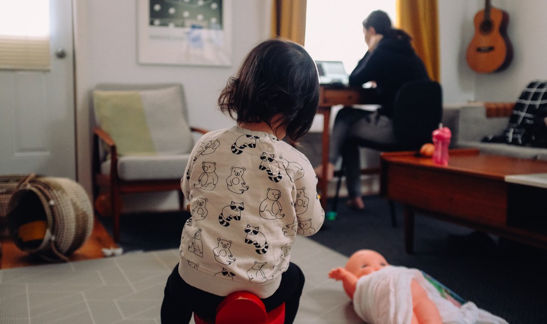 Kind im Vordergrund, Mutter arbeitet im Hintergrund am Computer.