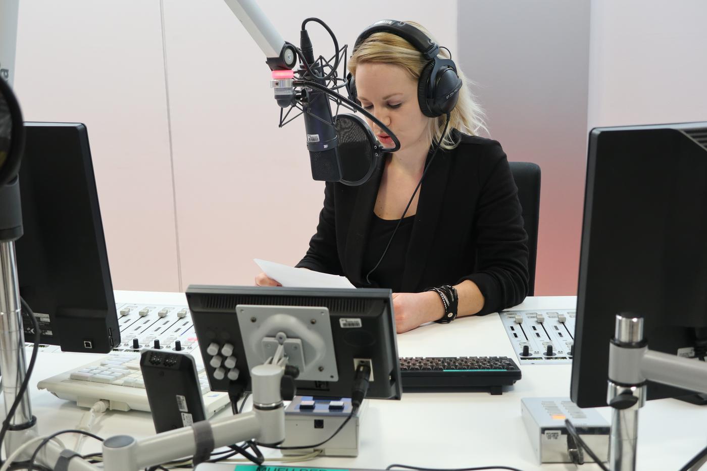 Isabelle befindet sich im Radiostudio und sitzt vor dem Mikrofon. In der Hand hält sie einen Zettel, von welchem sie den Text abliest.