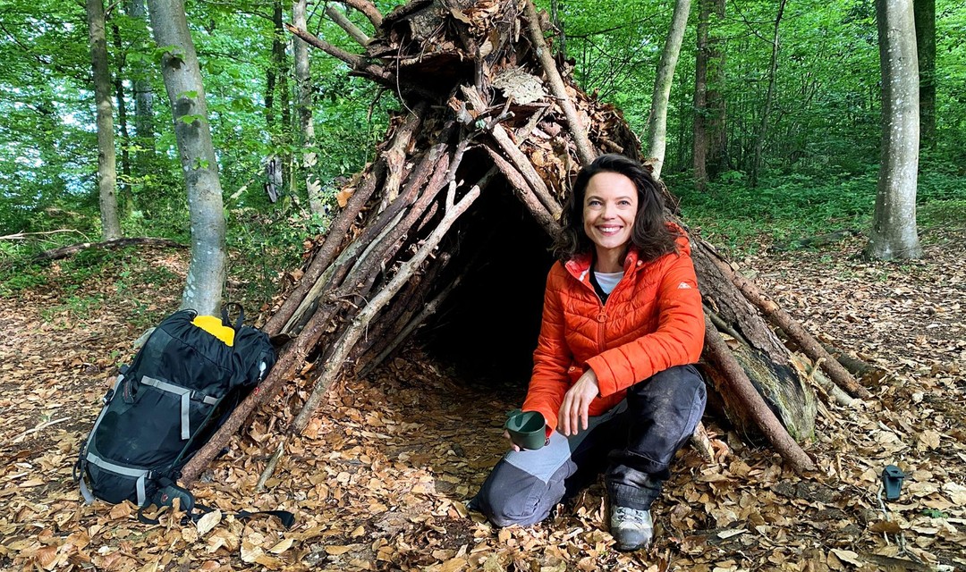 Mona Vetsch kniet vor einer selbst gebauten Hütte im Wald.