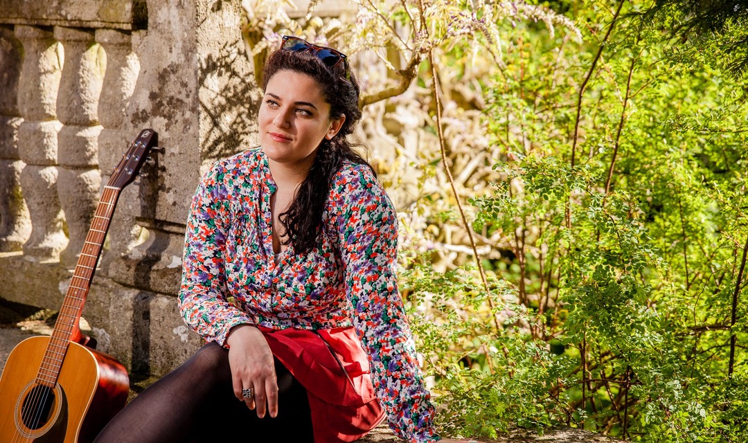 Rebekka Lindauer Frau mit schwarzen Locken und Blumenkleid in der Natur mit Gitarre neben sich