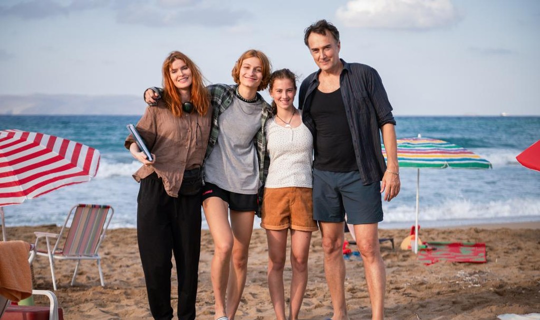 Das Foto zeigt ein Porträt der Patchworkfamilie am Strand. Links stehen Mutter Monica und Tochter Valeska, rechts anschliessend stehen Lena und ihr Vater Jacques. Im Hintergrund befinden sich das Meer, einige bunte Sonnenschirme und Badetücher sowie ein Camping-Klappstuhl.