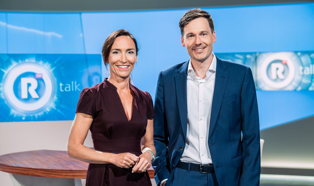 Die Moderatoren Nicole Frank (links) und Dominik Meier (rechts) stehen im Rundschau Talk Studio. Sie trägt ein weinrotes Kleid, er trägt einen dunkel blauen Anzug mit einem hell blauen Hemd.