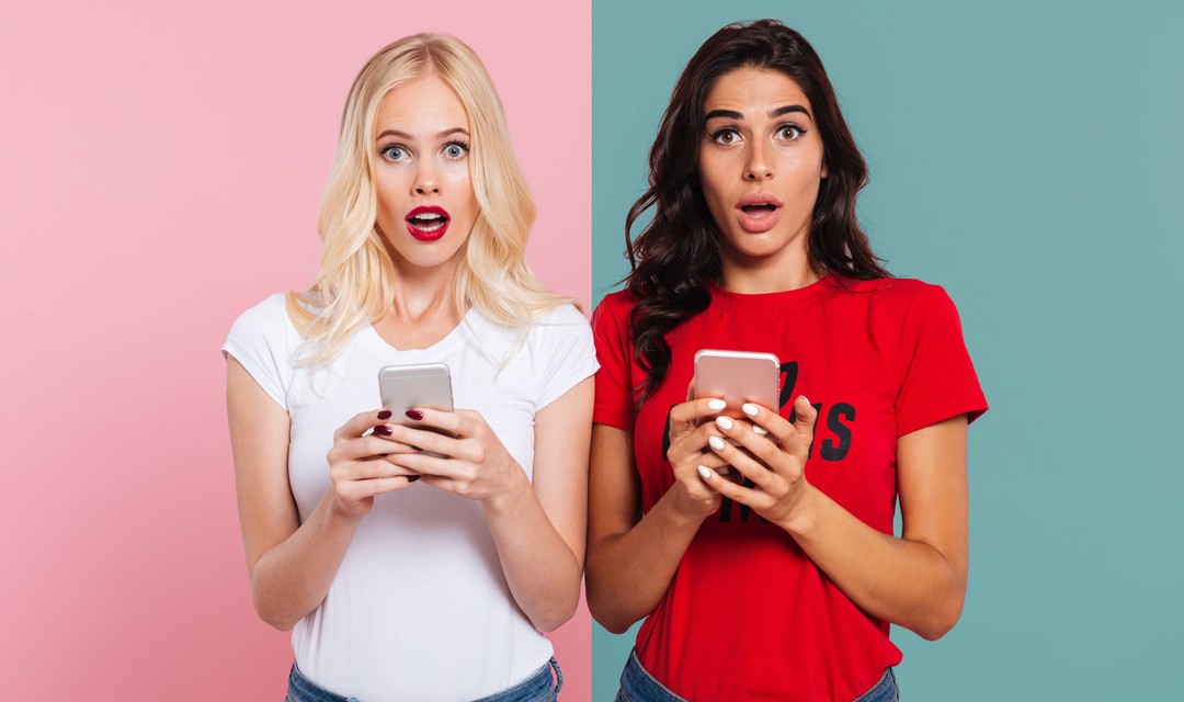 Zwei Frauen halten ihr Mobiltelefon in der Hand und blicken verwundert in die Kamera