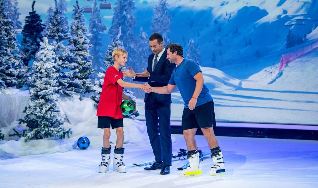 Das Foto zeigt Beat Feuz, den Buben Matti und Moderator Kai Pflaume. Matti und Beat Feuz tragen T-Shirt, kurze Hosen und Skischuhe. Sie geben sich lächelnd die Hand. Kai Pflaume steht zwischen den beiden. Matti hält einen grünen Fussball in der linken Hand. Alle Personen stehen im Studio. Hintergrund und Studiodekoration zeigen verschneite Tannen und eine verschneite Berglandschaft.