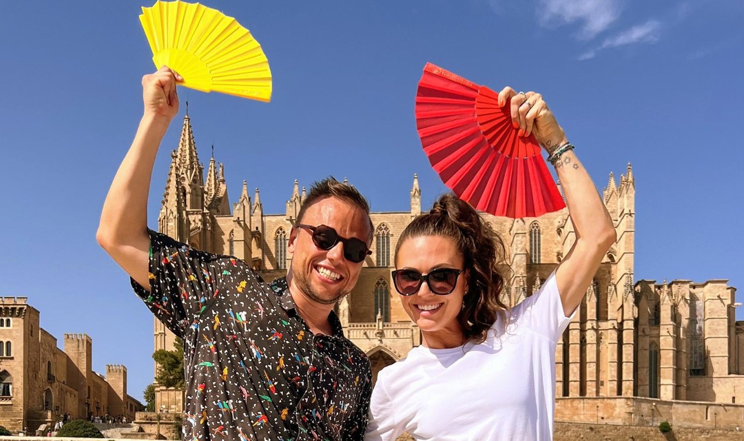 Das Foto zeigt die beiden Moderierenden Vila Tami und Jan Fitze vor einer Kathedrale auf Mallorca in tänzerischer Pose. Sie strecken beide einen geöffneten Fächer in die Höhe.