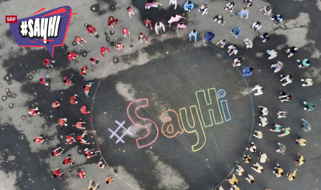 Drohnenaufnahme von Schülerinnen und Schüler beim Tanzen auf dem Pausenplatz. Alle sind um einen grossen Kreis verteilt. In der Mitte des Kreises steht bunt mit Kreide geschrieben «#SayHi»
