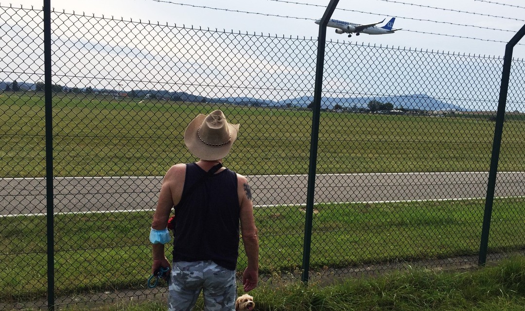 Edi steht vor einem Zaun und beobachtet ein vorbeifliegendes Flugzeug