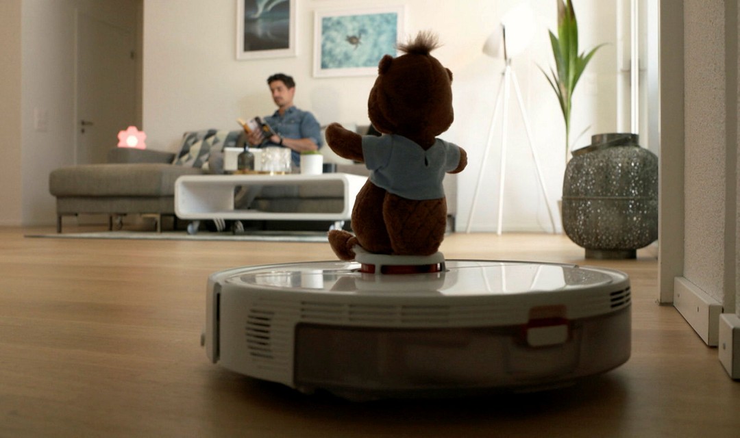 Ein Staubsauger-Roboter steht im Vordergrund, Tobias Müller sitzt im Hintergrund auf der Couch.