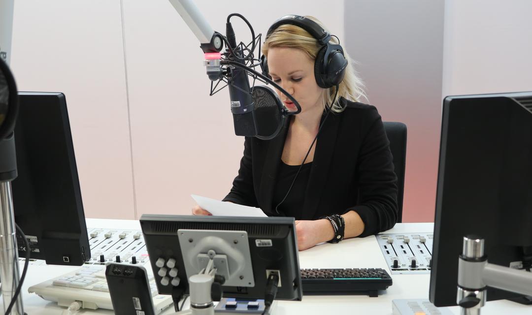 Podcast-Autorin steht im Studio vor dem Mikrofon und liest von einem Blatt Papier ab
