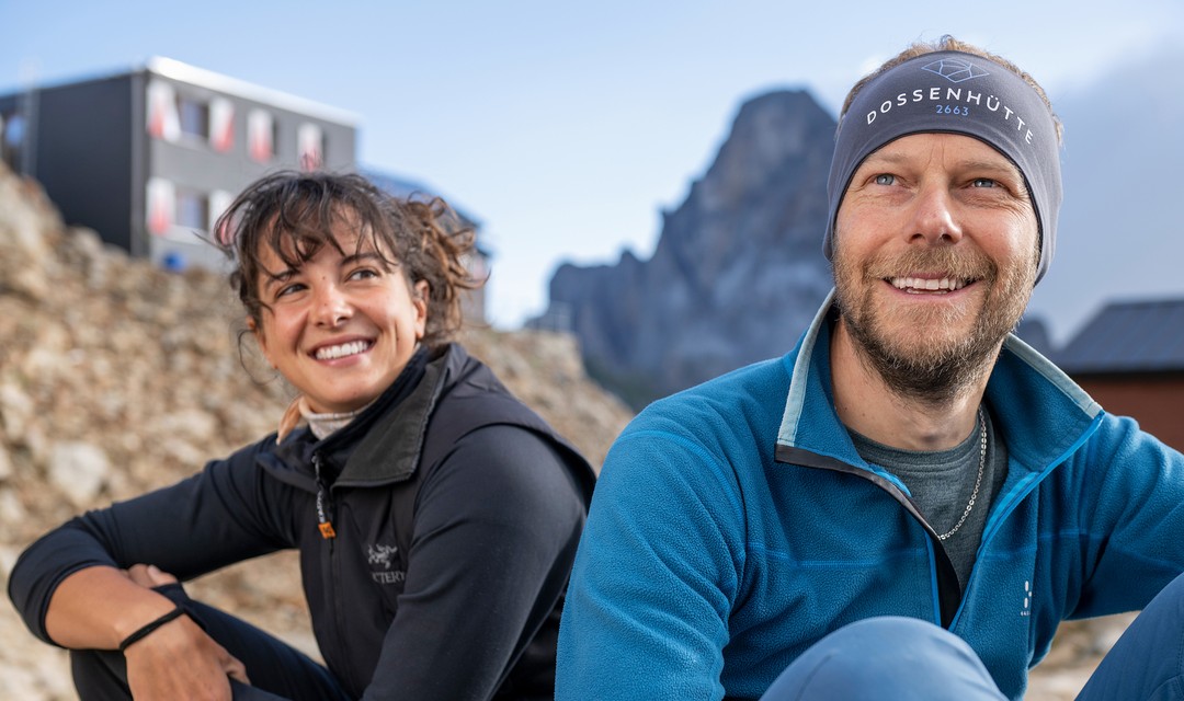 Hüttenhilfe Ronja Trachsler und Hüttenwart David Zweifel sitzen draussen. Im Hintergrund eine Bergspitze und die Dossenhütte