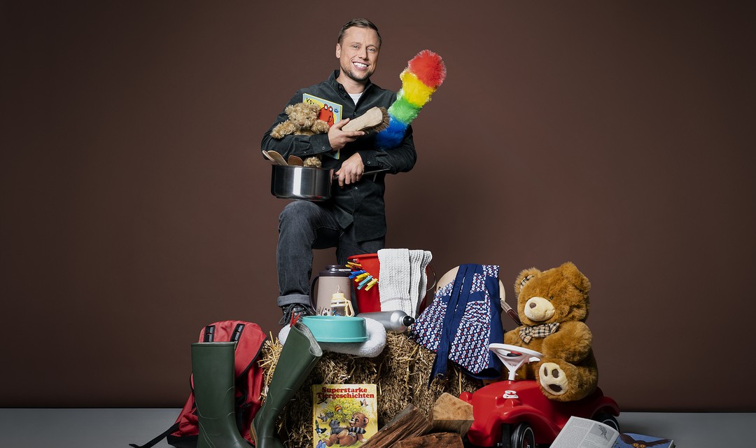 Foto von Jan Fitze in mitten unterschiedlichster Gegenstände wie Gummistiefel, Teddybären, Pfannen und Putzutensilien