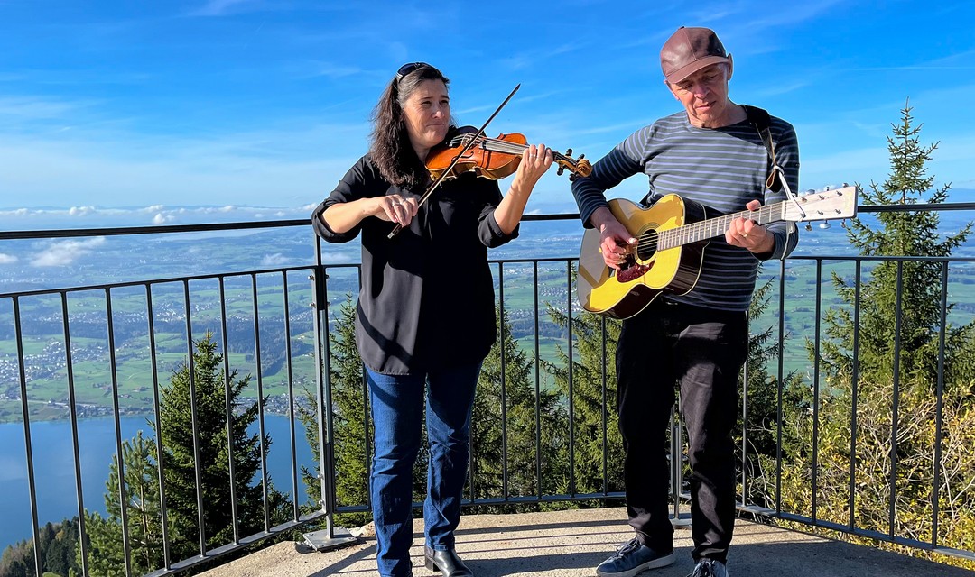 Foto: Eine Frau und ein Mann musizieren auf der Rigi mit der Aussicht über den See im Hintergrund. Die Frau spielt Geige, der Mann Gitarre