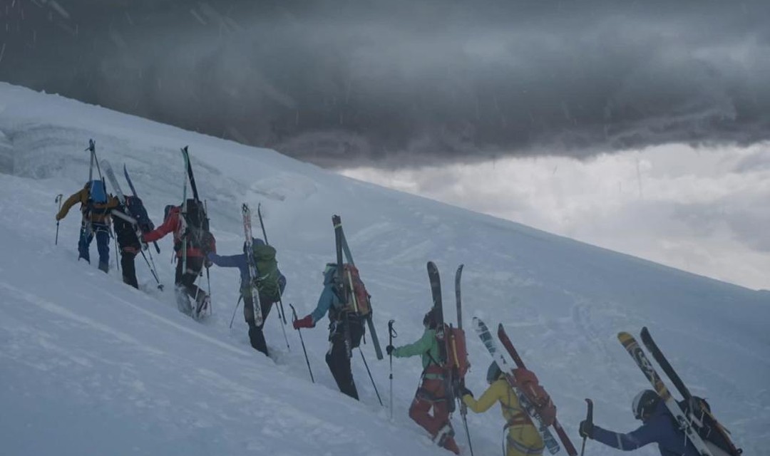 Das Foto zeigt eine Gruppe von 8 Tourengänger:innen, die hintereinander einen schneebedeckten Hang hinauflaufen. Am Rücken tragen sie Rucksack und Skier. Über ihnen ist eine dunkle Wolkenfront.