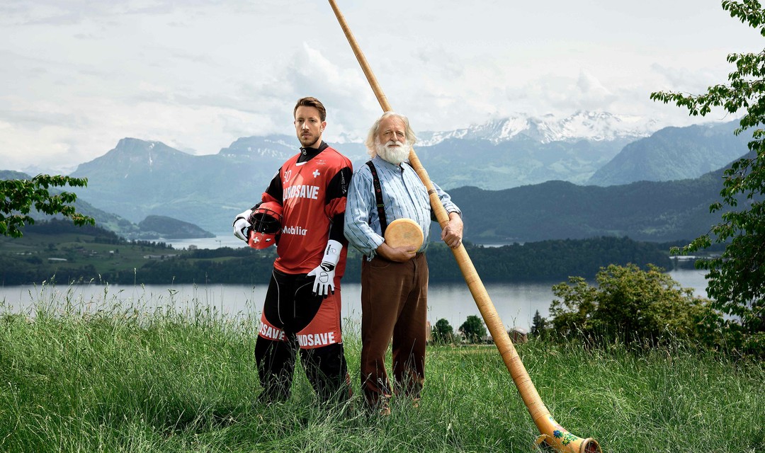 Pascal Meier, Torwart der Schweizer Unihockey Nationalmannschaft, posiert mit einem Alphornbläser vor einem See. Im Hintergrund sind Berge zu sehen.