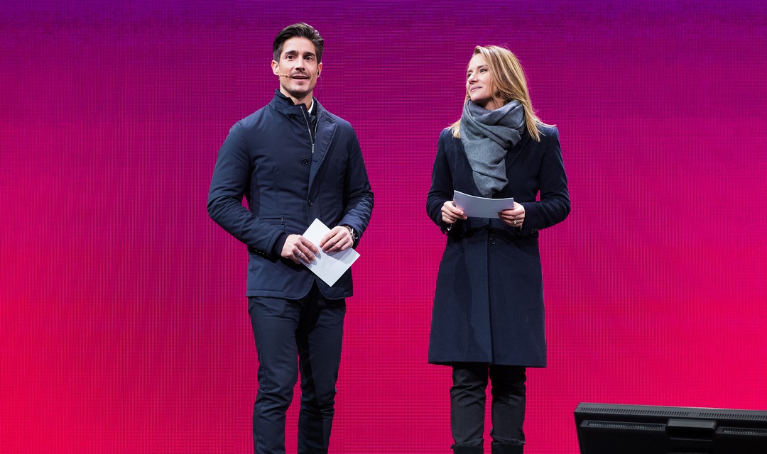 Moderator Tobias Müller und Moderatorin Wasiliki Goutziomitros auf einer Bühne vor pinkem Hintergrund
