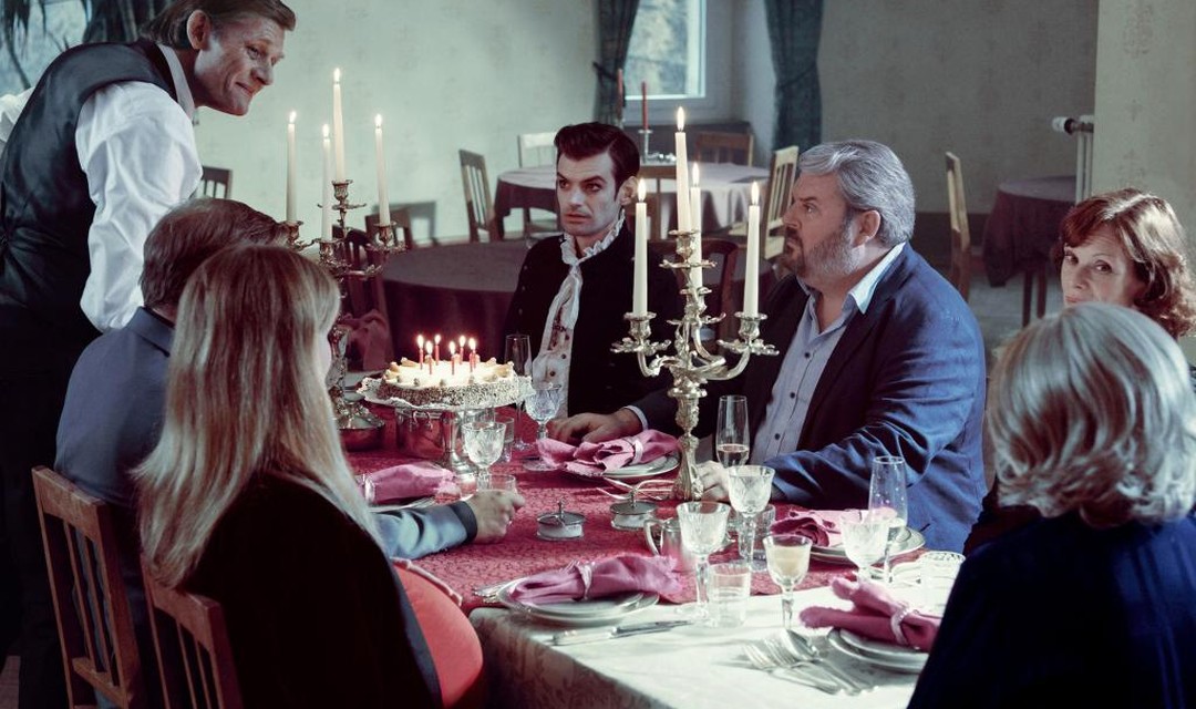 Das Foto zeigt mehrer Menschen an einem festlich gedeckten Tisch. Auf den Tischen stehen Kristallgläser und silberne Kerzenständer sowie eine Rüeblitorte mit brennenden Kerzen. Zu den Gästen gehören die Figuren Fabio Testi und Luc Conrad.