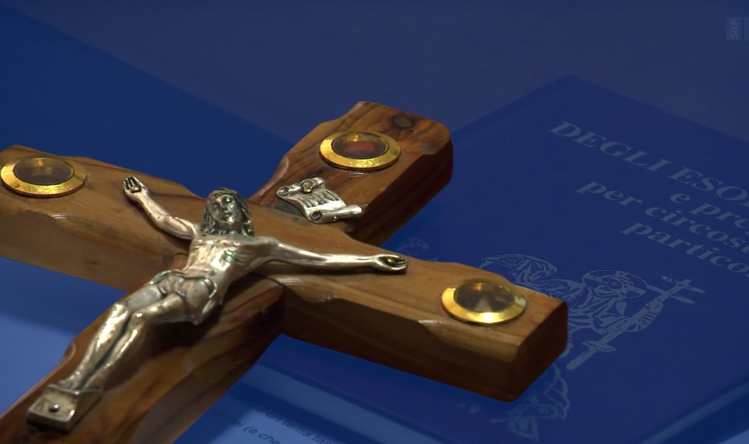 Das Foto zeigt ein Holzkreuz. Die Jesus-Figur darauf ist aus Metall. Das Kreuz liegt auf einem italienischen Gebetsbuch für Exorzismen.