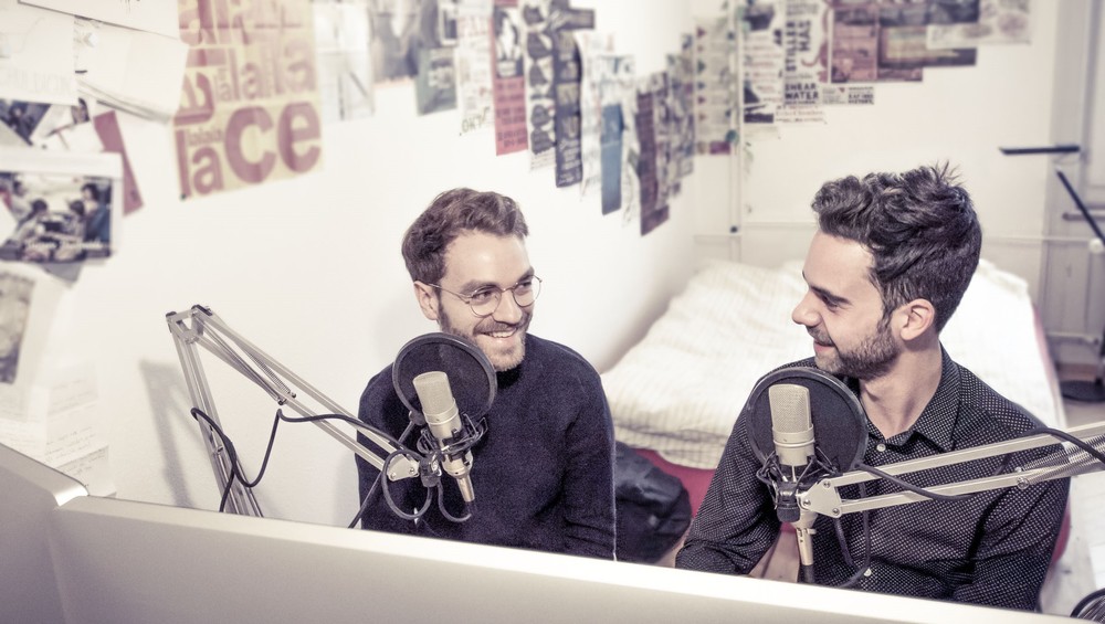 Zwei junge Herren sitzen in einem Schlafzimmer hinter Mikrophonen
