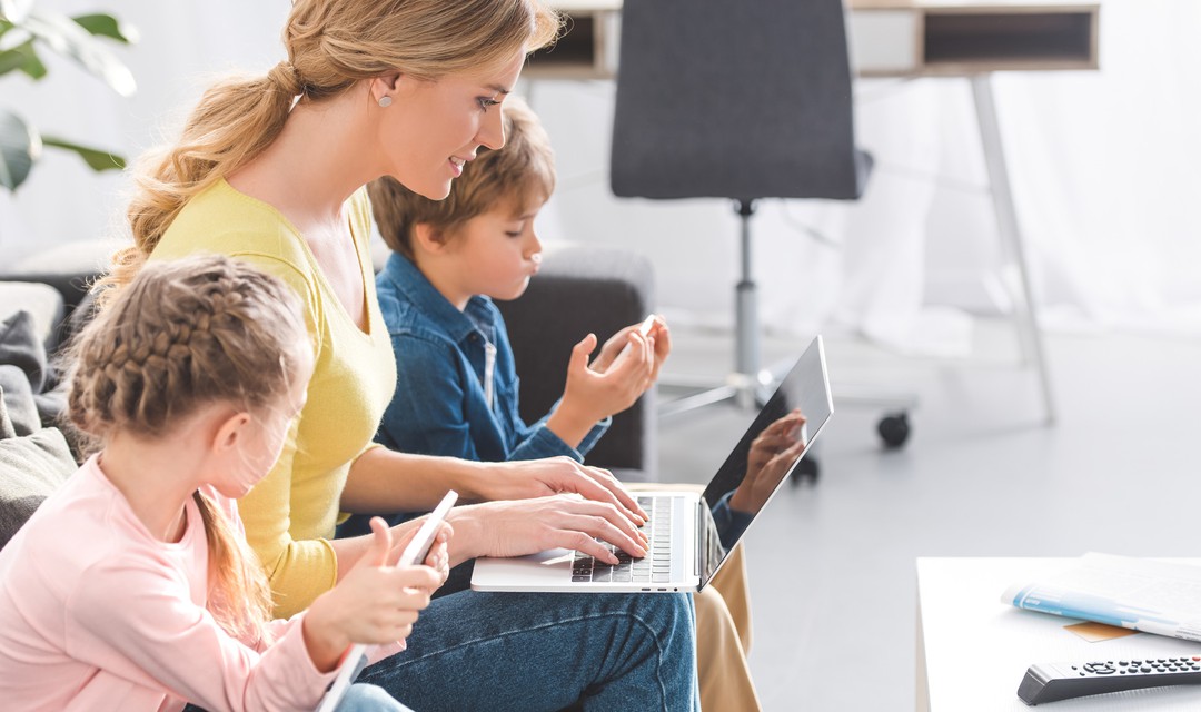 Eine Frau sitzt mit ihren beiden Kindern im Wohnzimmer und zeigt ihnen was auf dem Laptop