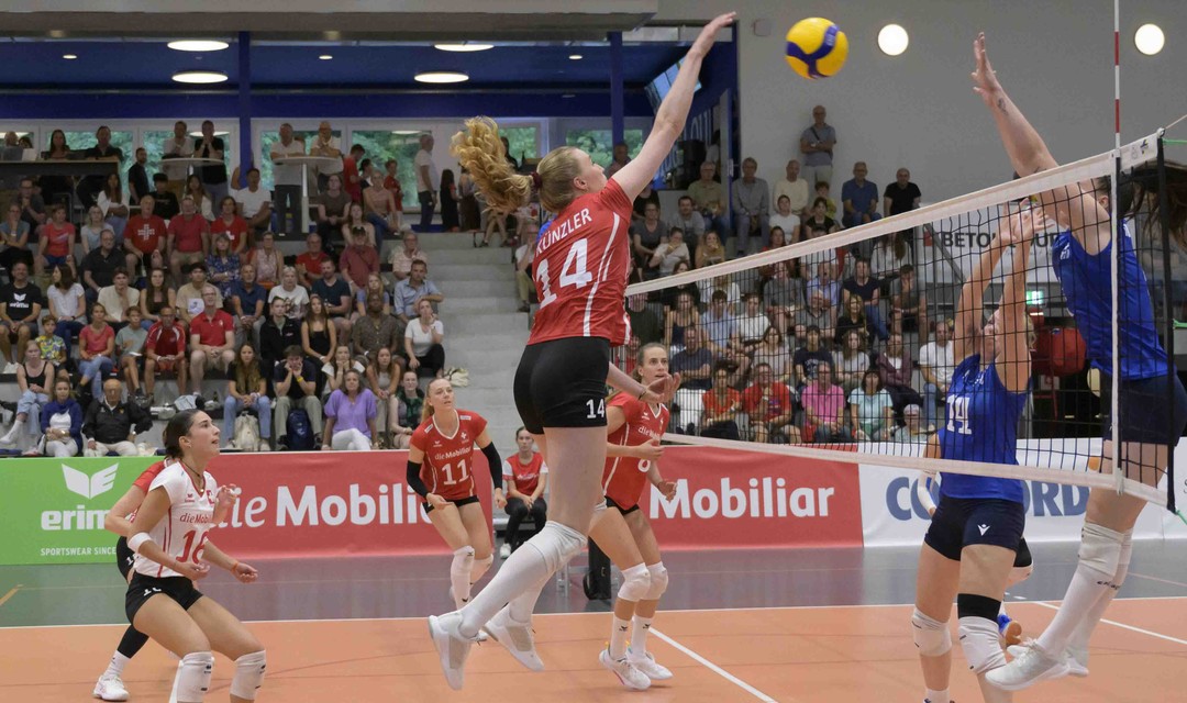 Das Foto zeigt eine Frauen-Volleyball-Mannschaft während eines Spiels. Laura Künzler springt am Netz hoch und schlägt den Ball auf die gegnerische Seite.