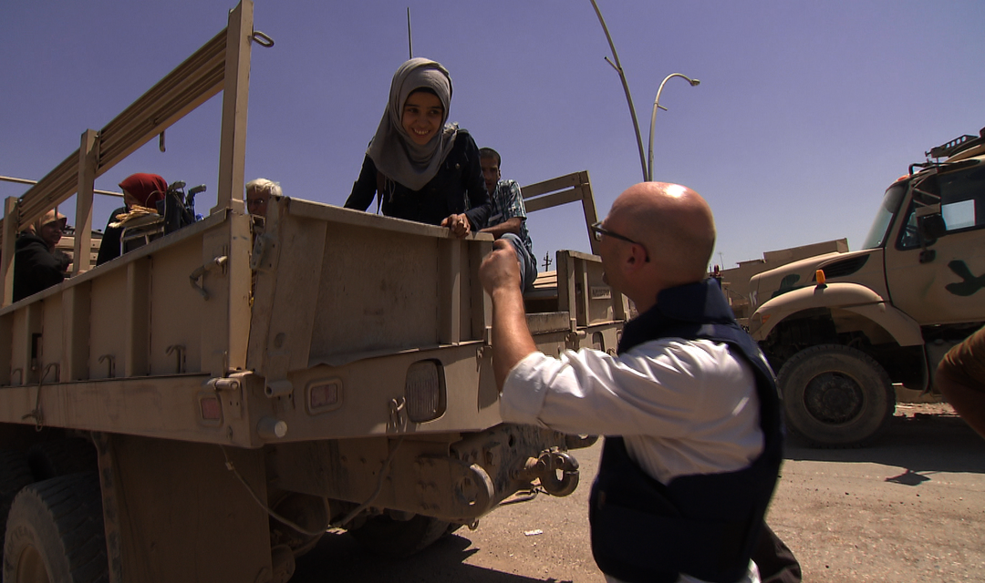 Pascal Weber ist mit schusssicherer Weste im Einsatz. Er reicht einem Mädchen die Hand, im Hintergrund sind Militärfahrzeuge zu sehen.