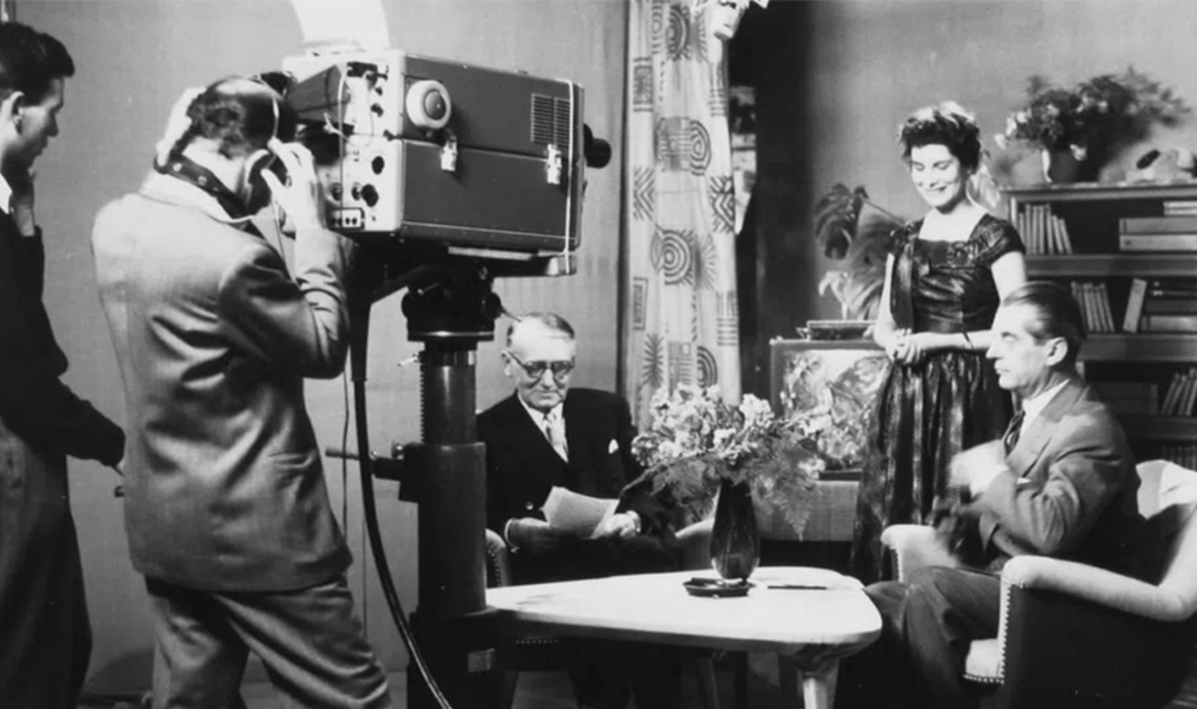 Schwarz-Weiss Archivbild Kameramann filmt Szene zwei Männer sitzen am Tisch eine Frau steht
