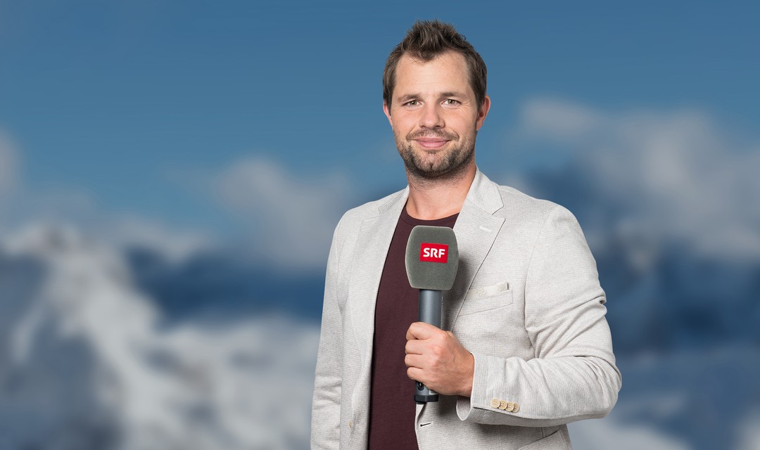 Das Foto zeigt ein Porträtbild von Beat Feuz. Er hält ein SRF-Mikrofon in der linken Hand und schaut schmunzelnd in die Kamera. Im Hintergrund ist eine sonnige, schneebedeckte Berglandschaft verschwommen wahrnehmbar.
