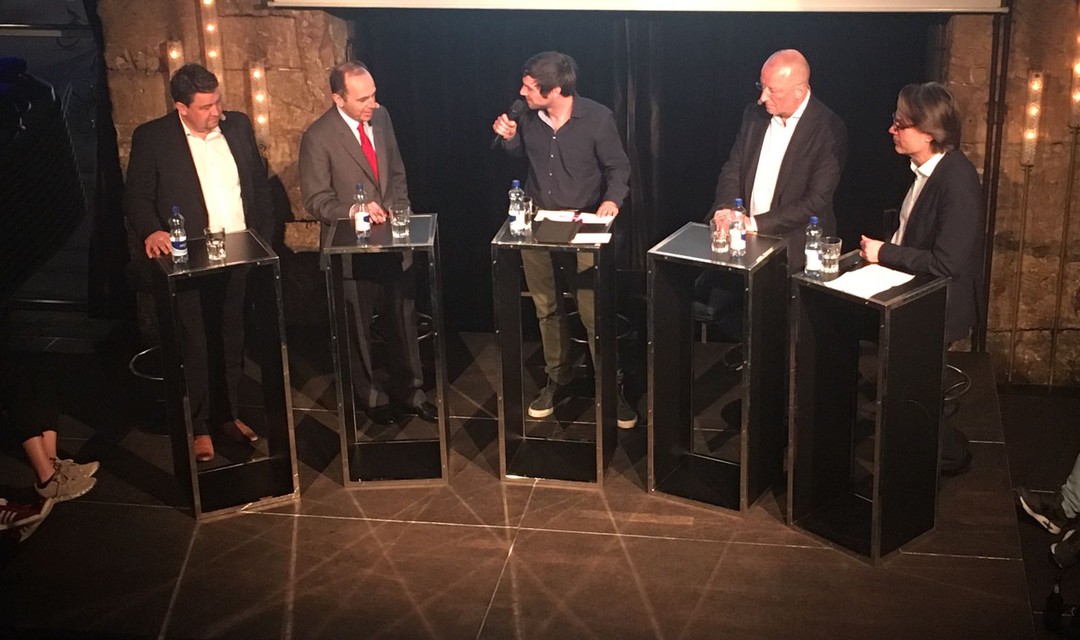 Vier Herren auf Podium am Diskutieren und ein Moderator der Mikrofon in der Hand haelt