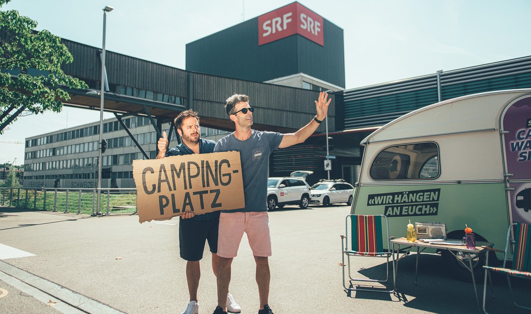 Die Moderatoren Philippe Gerber und Marco Thommann stehen winkend vor einer Caravane.
