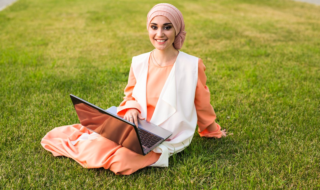 Junge Muslimin im Gras sitzend mit Laptop