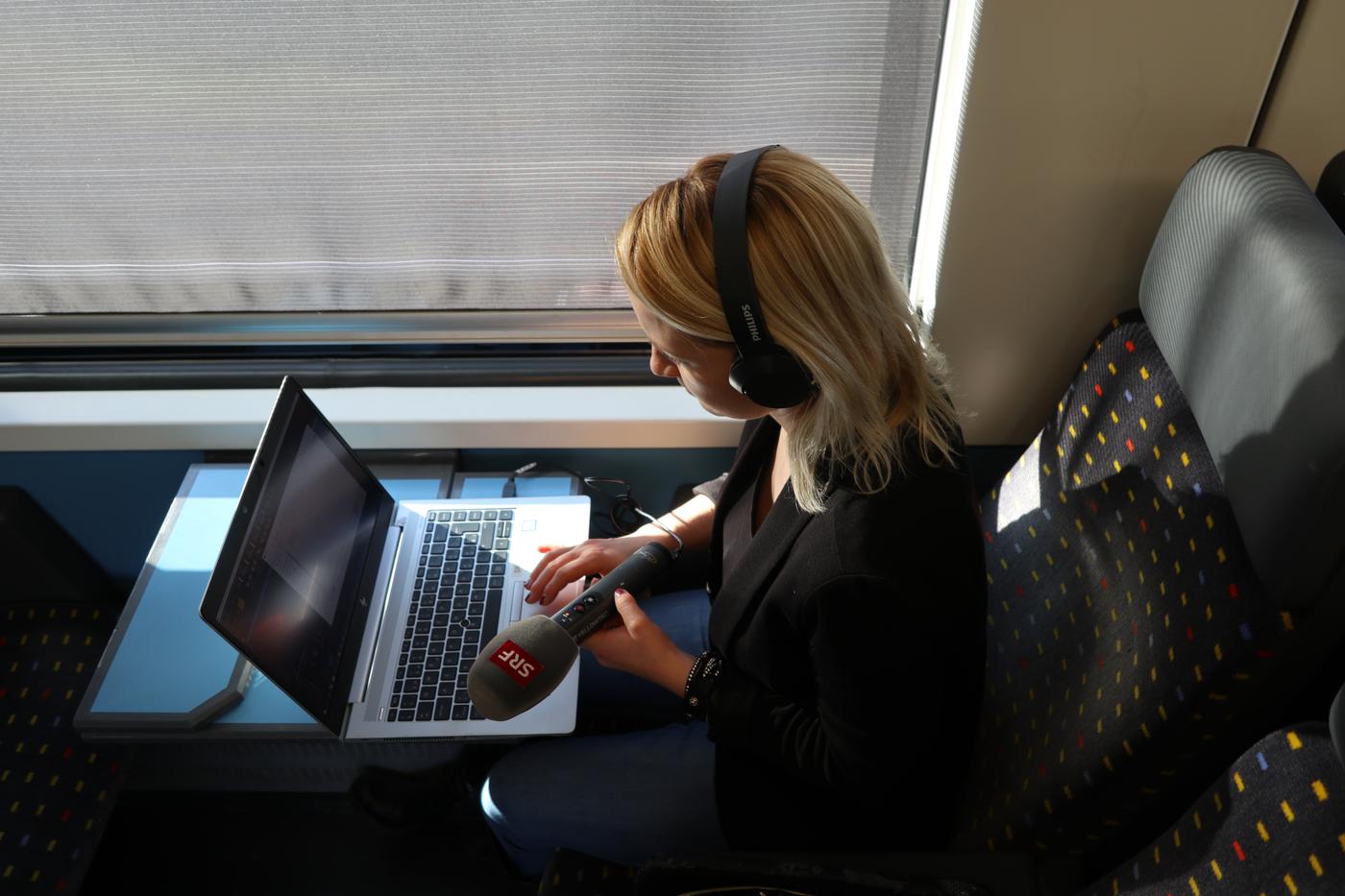 Isabelle sitzt mit Kopfhörern in einem Zugabteil und hat den Laptop vor sich auf den Tisch gestellt