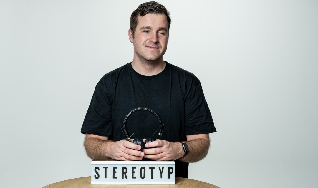 Yves Kilchör steht vor einem Tisch, auf welchem ein «Stereotyp»-Schild und Kopfhörer liegen