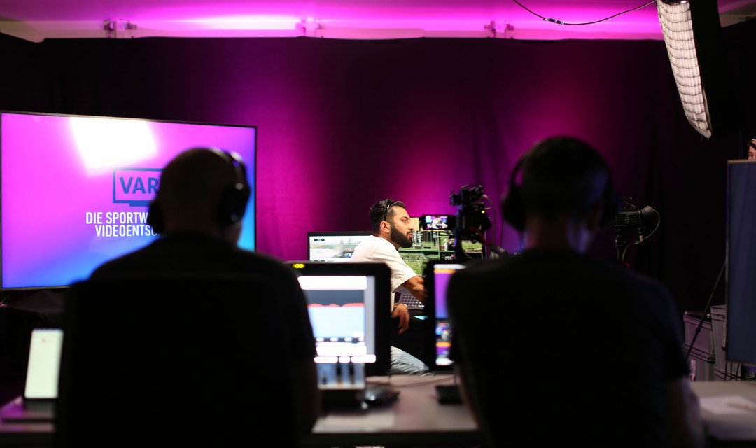 Zwei Personen sitzen an einer Regie, im Hintergrund ist ein Moderator zu sehen.