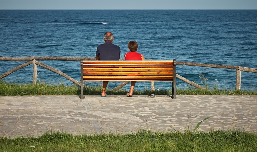 Grossvater und Enkel sitzen auf einer Bank und blicken ins offene Meer